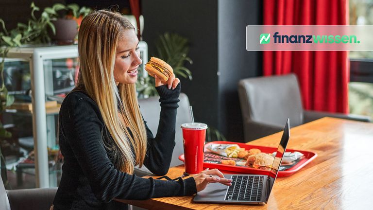 Blonde Frau isst in einem Fast-Food-Restaurant und arbeitet gleichzeitig am Laptop.