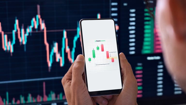 Trader blickt auf sein Handy, das eine Bearish Engulfing Kerzenformation anzeigt. Im Hintergrund ein Trading Monitor.