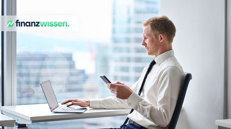 Blonder Trader arbeitet in einem Hochhaus-Büro an seinem Laptop und vergleicht Daten mit seinem Smartphone